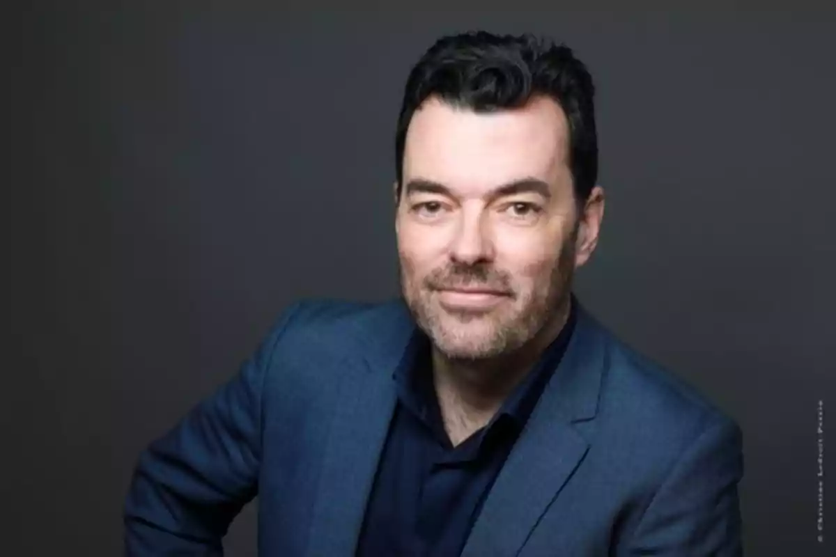 Un hombre con barba y cabello oscuro lleva un traje azul y camisa oscura posando frente a un fondo gris.