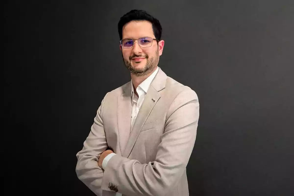 Hombre de cabello oscuro con gafas y barba corta usando saco beige y camisa blanca posando con los brazos cruzados frente a un fondo gris oscuro