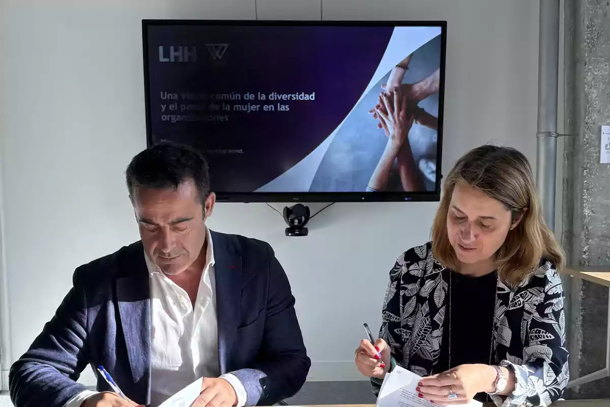 Dos personas firmando documentos en una oficina con una pantalla al fondo que muestra una presentación sobre diversidad y el papel de la mujer en las organizaciones