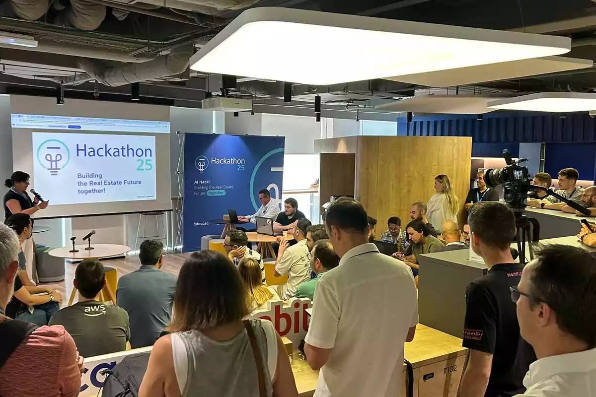 Personas participando en un hackathon de tecnología inmobiliaria, con una presentación proyectada y varias personas observando y trabajando en computadoras.