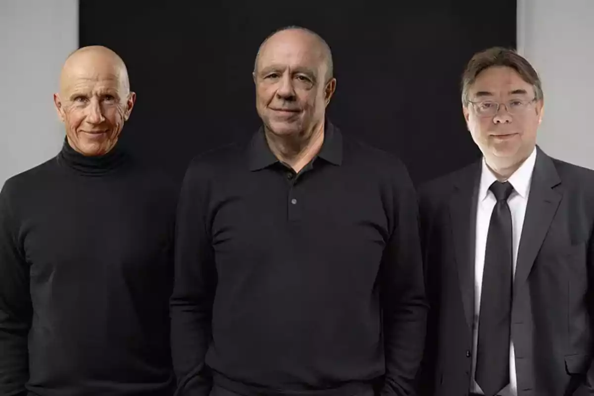Tres hombres posan juntos frente a un fondo oscuro, dos de ellos visten ropa negra informal y el tercero lleva traje y corbata.