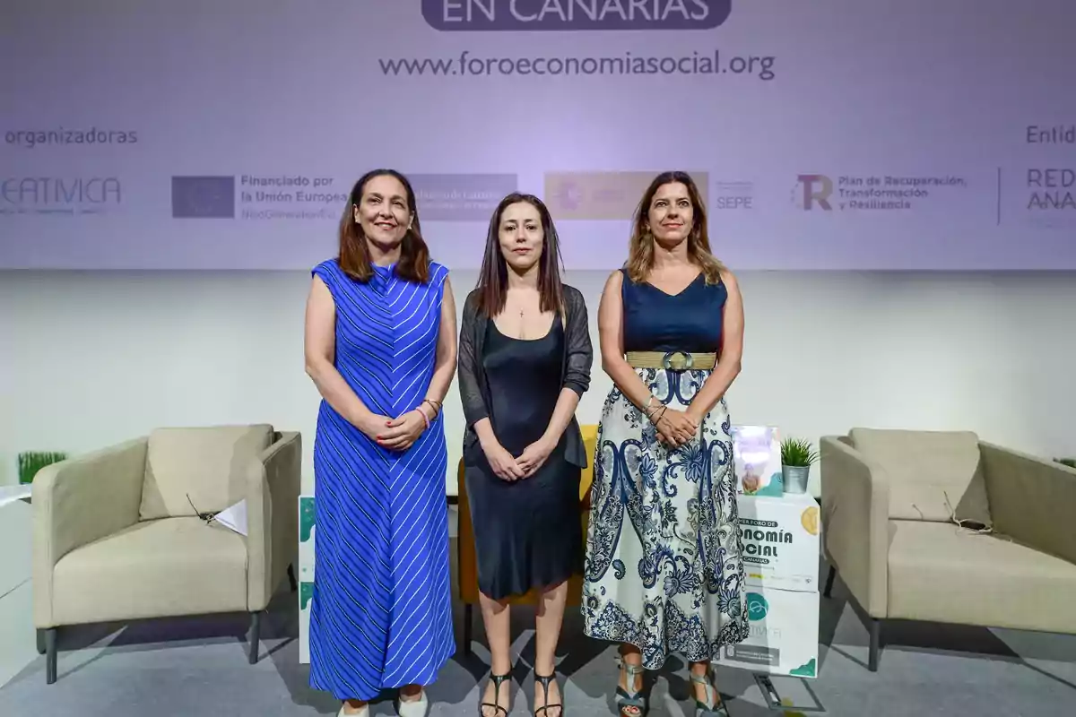 Tres mujeres posan de pie en un escenario durante un evento de economía social en Canarias, con sillones y carteles de fondo.
