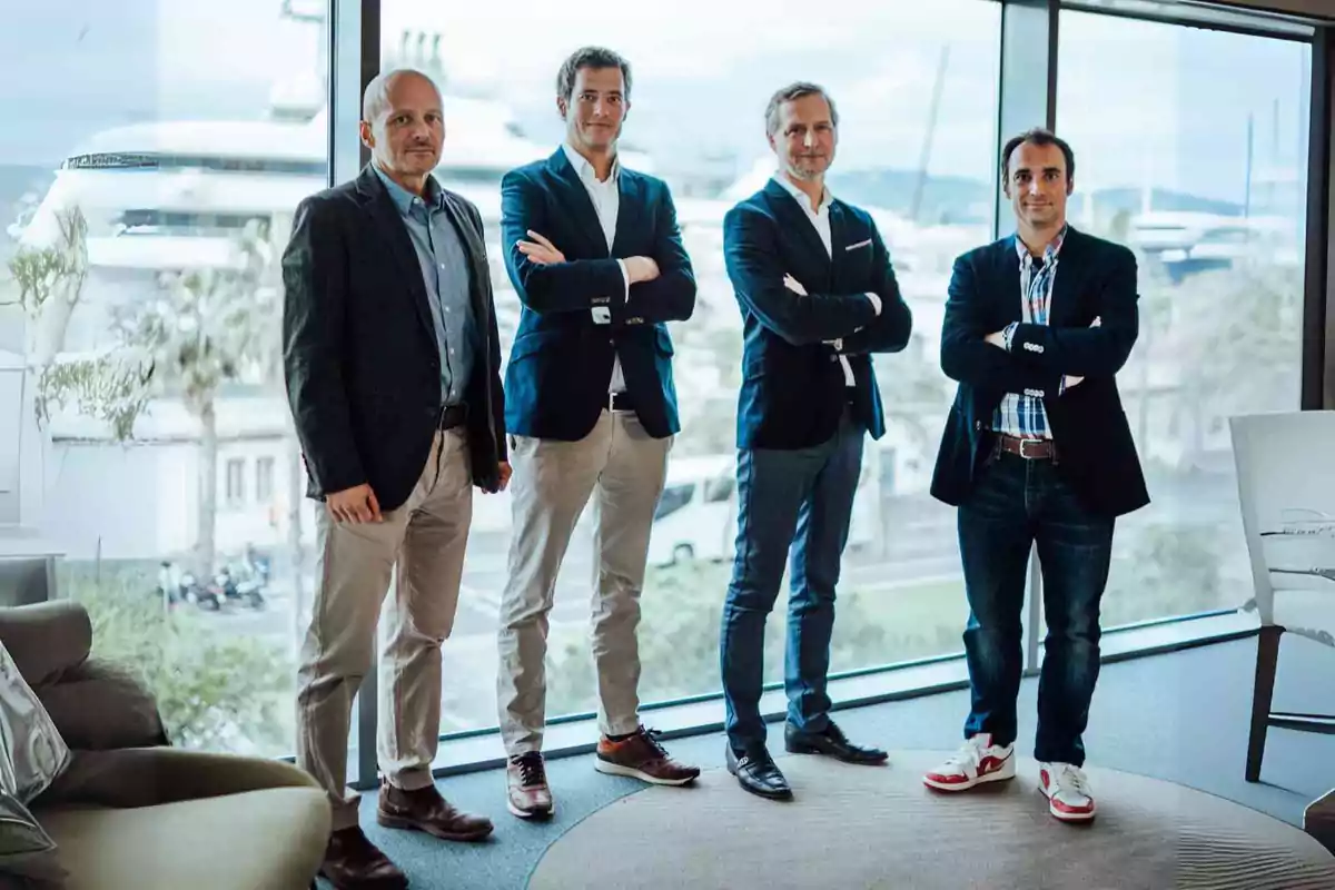 Cuatro hombres de pie en una oficina moderna con ventanales grandes, todos vestidos con saco y pantalón, posando juntos y mirando a la cámara