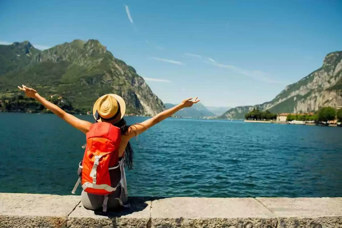 Mujer con mochila y sombrero sentada frente a un lago rodeado de montañas con los brazos extendidos disfrutando del paisaje