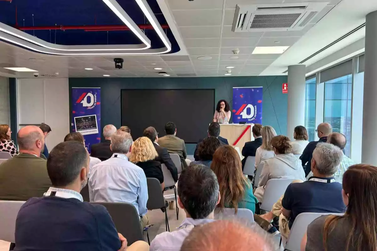 Personas sentadas en una sala de conferencias escuchando a una mujer que habla en un podio frente a una pantalla y dos carteles con el número 40