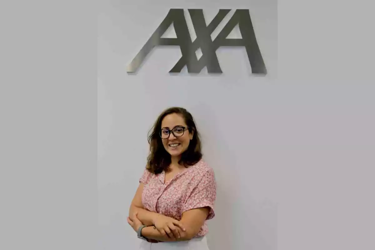 Mujer con gafas y camisa rosa sonríe frente a una pared blanca con el logotipo de AXA en la parte superior