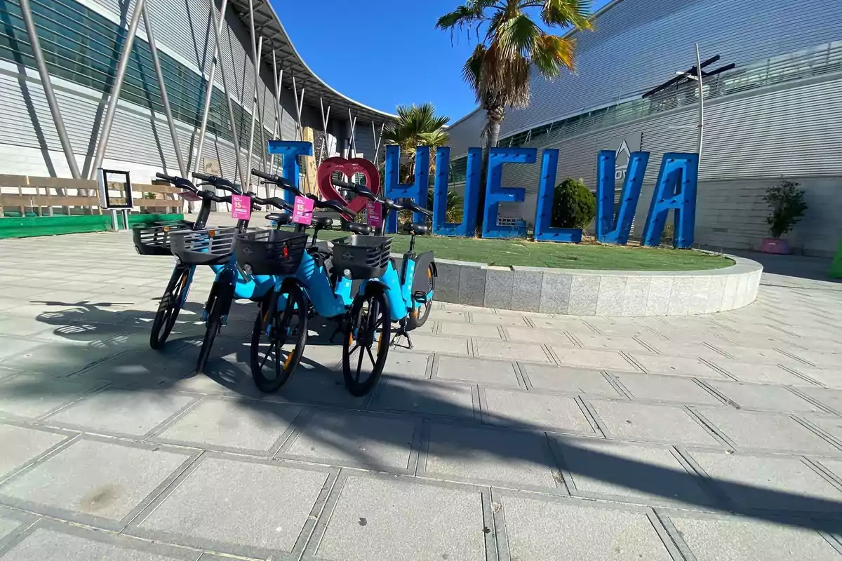 Bicicletas azules de alquiler estacionadas frente a un letrero grande que dice I love Huelva junto a una palmera y un edificio moderno