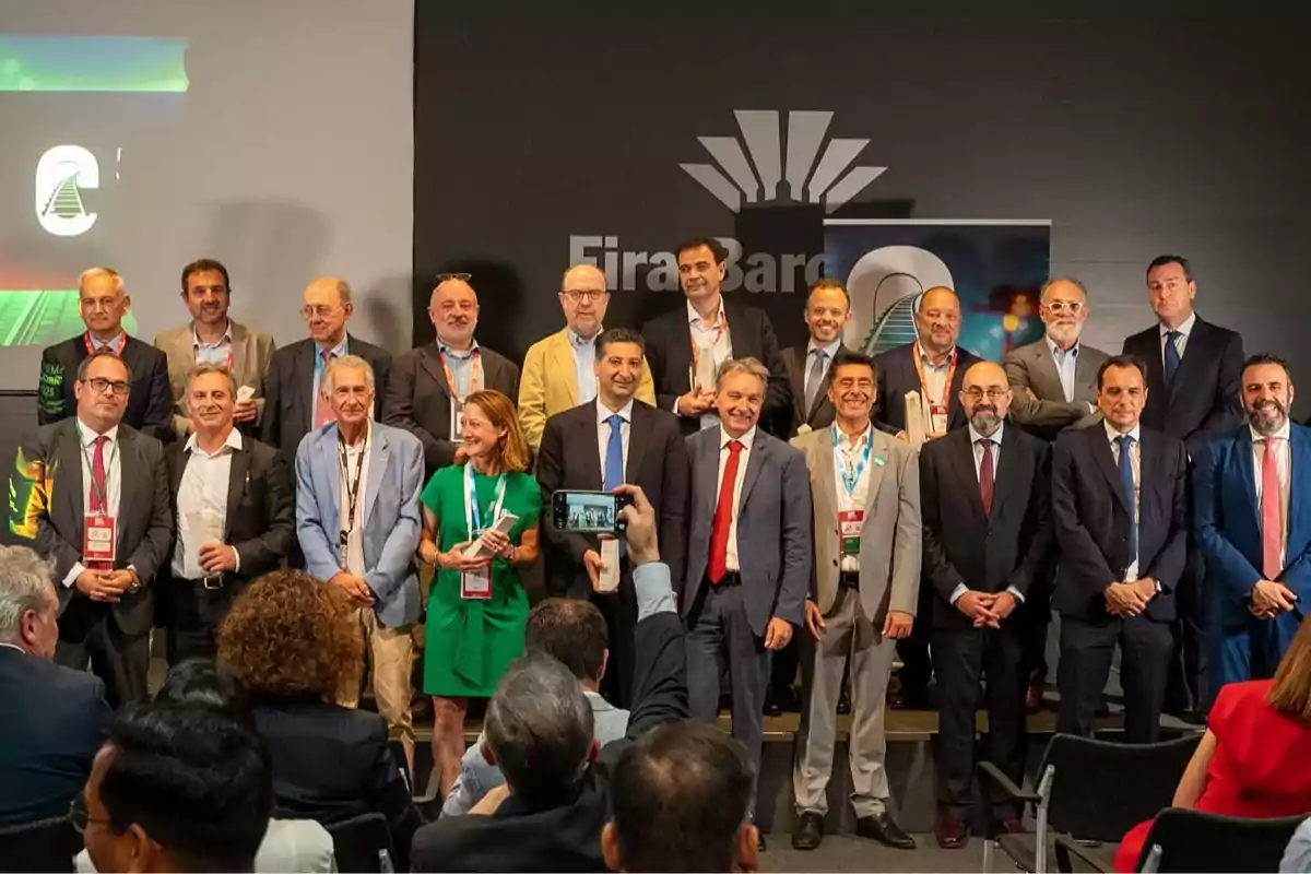 Un grupo de personas posando para una foto en un evento profesional frente a un fondo con el logo de Fira Barcelona mientras una persona en el público toma una foto con su teléfono móvil