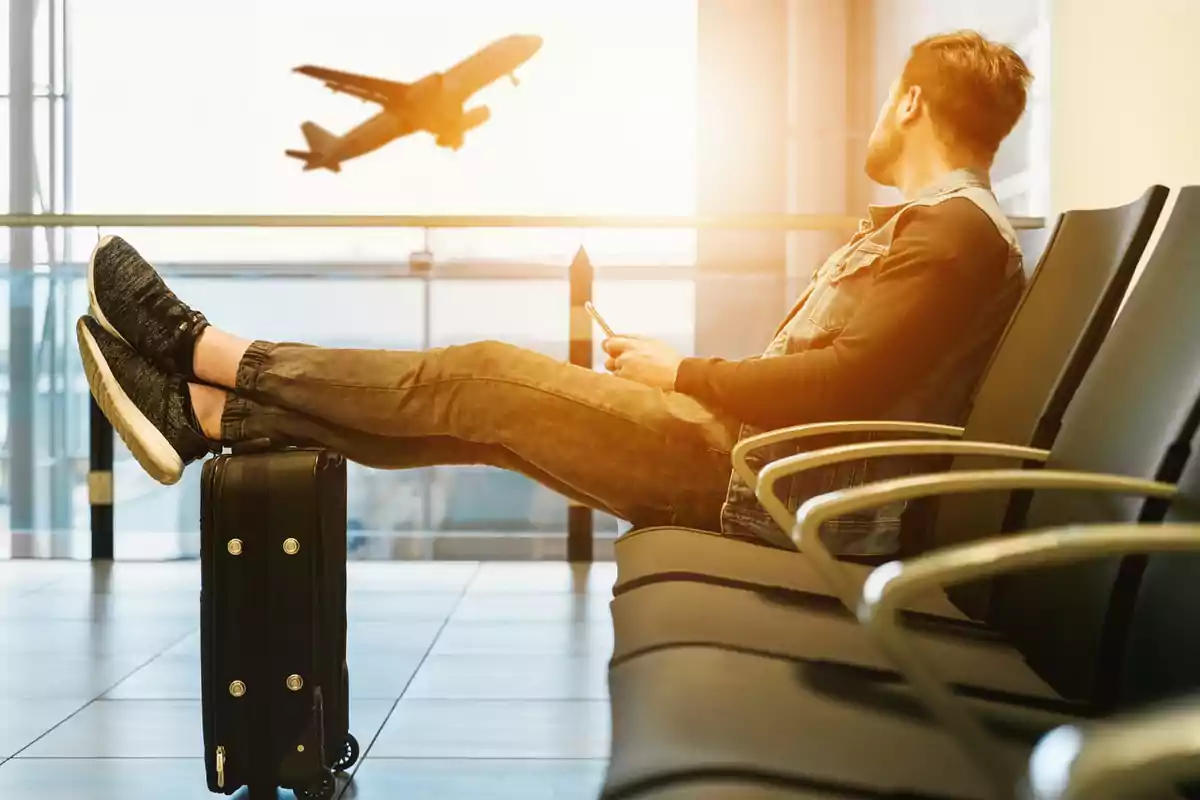 Hombre sentado en una sala de espera de aeropuerto con los pies sobre una maleta mirando un avión despegando por la ventana