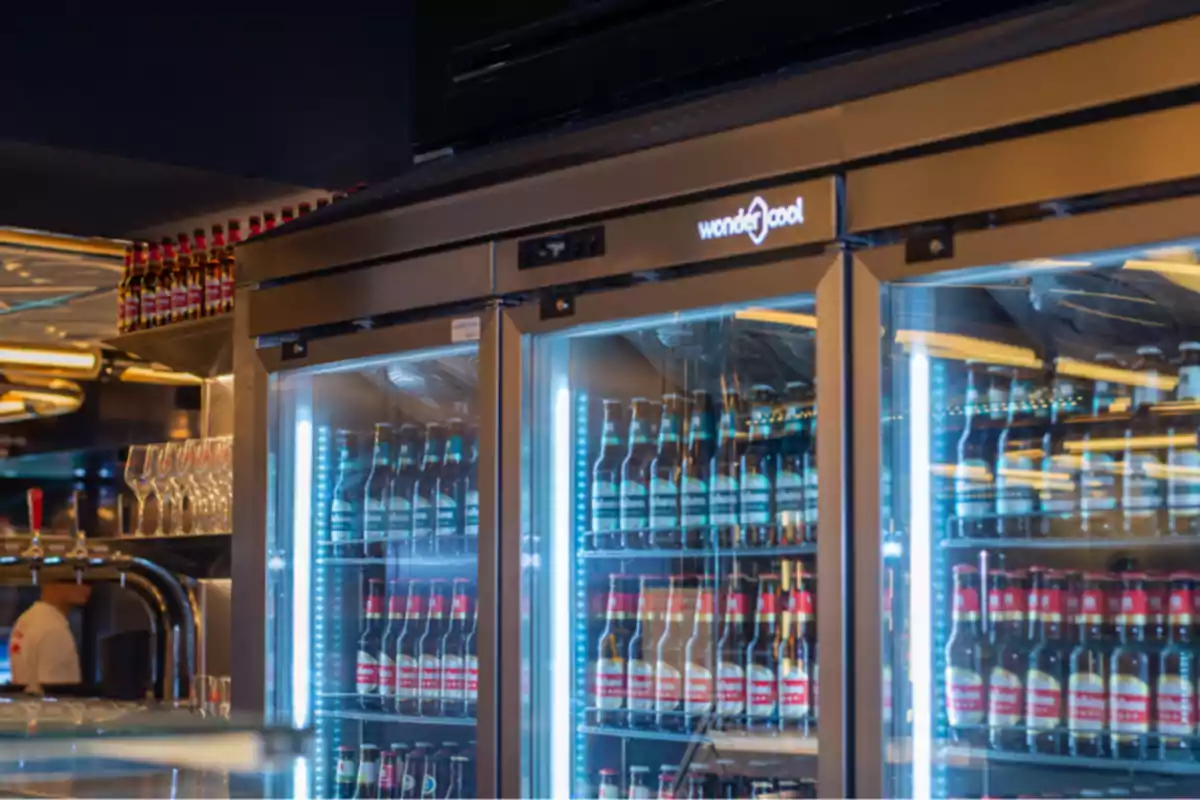 Refrigerador de bar con puertas de vidrio lleno de botellas de cerveza y copas de cristal al lado
