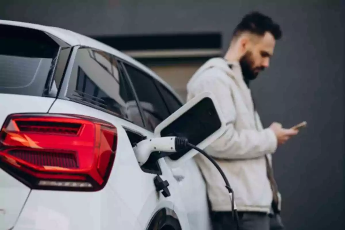 Hombre usando su teléfono móvil mientras un auto eléctrico blanco se está cargando