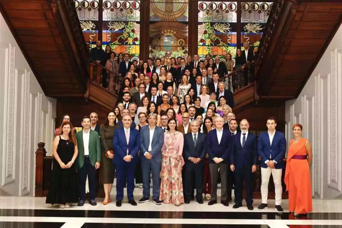 Un gran grupo de personas elegantemente vestidas posando en una escalera de madera con vitrales coloridos al fondo en un edificio de estilo clásico