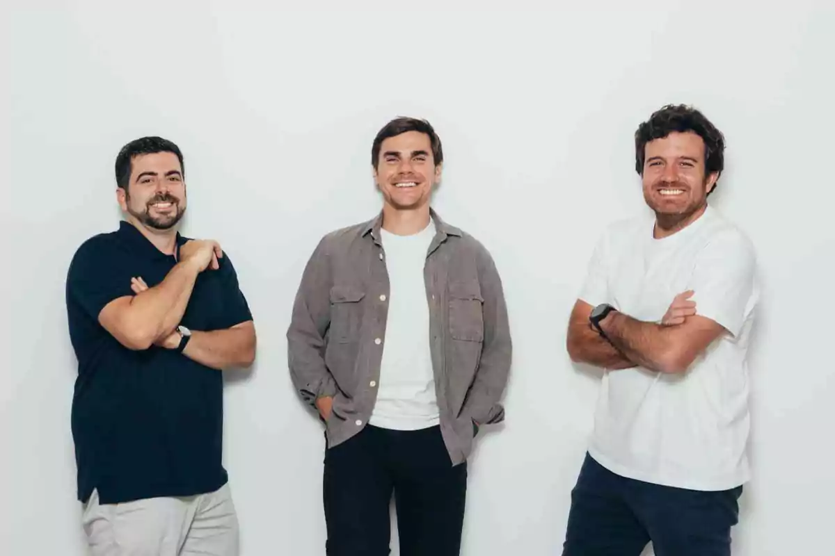 Tres hombres jóvenes sonríen y posan de pie frente a una pared blanca