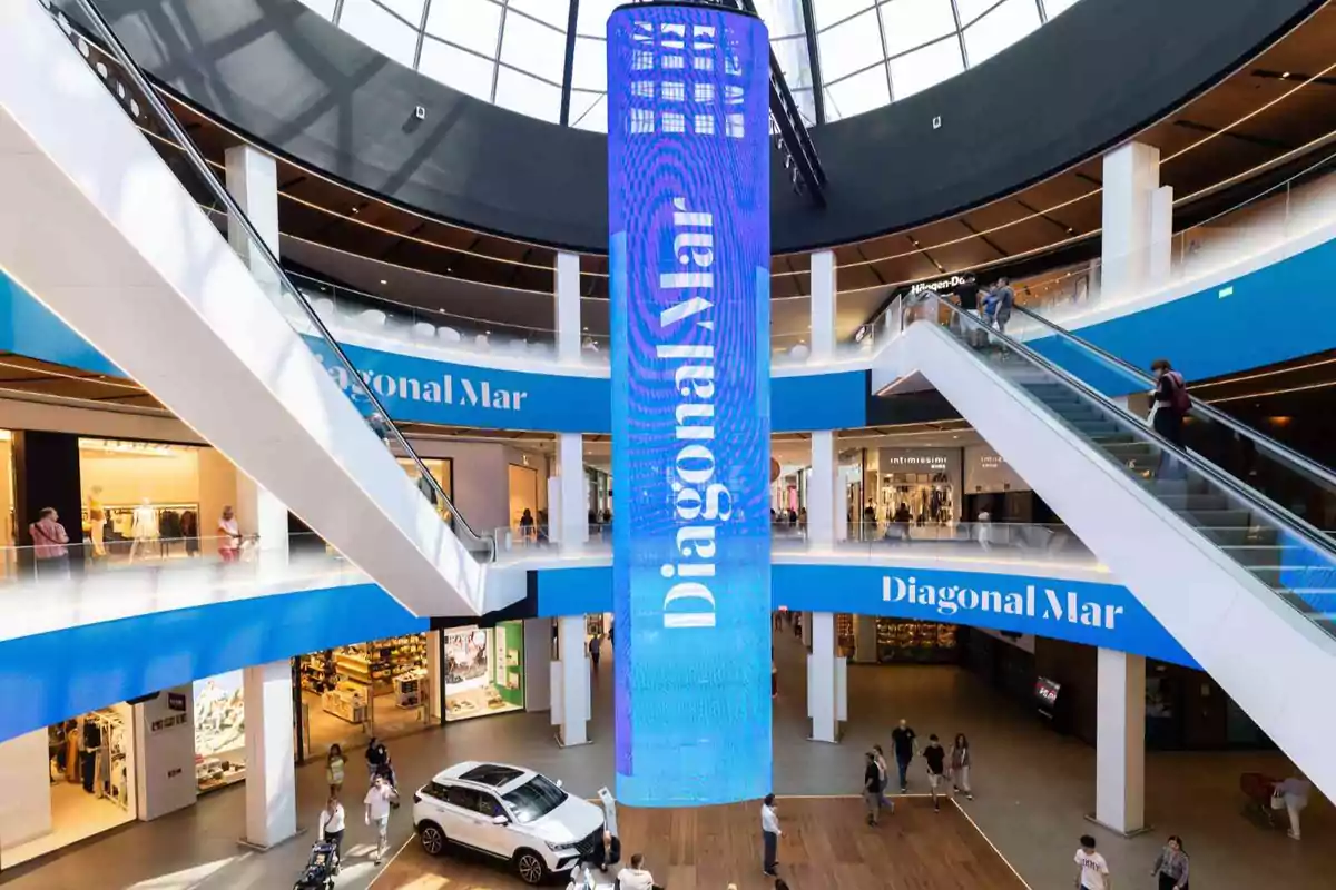 Centro comercial moderno con una gran pantalla azul vertical en el centro y personas caminando y usando escaleras eléctricas
