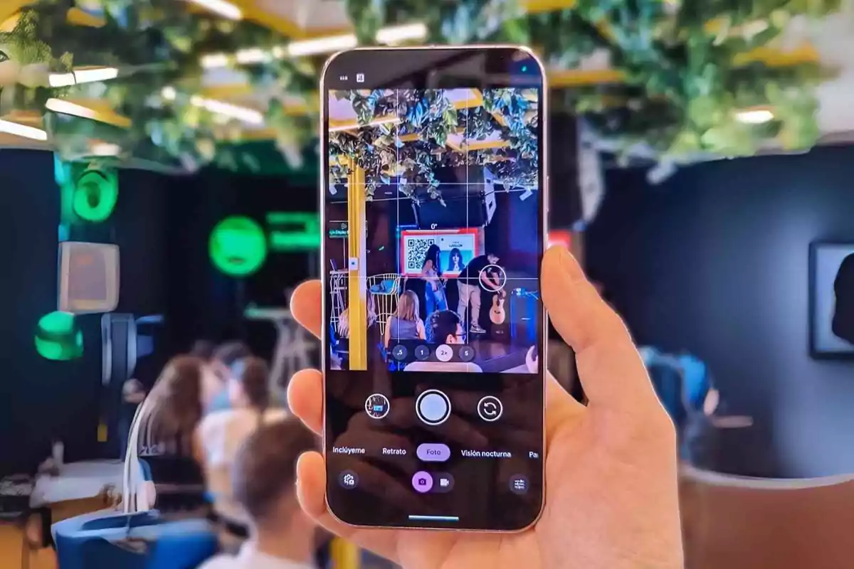 Persona sosteniendo un teléfono móvil que toma una foto de un evento en un espacio cerrado con varias personas sentadas y una pantalla al fondo