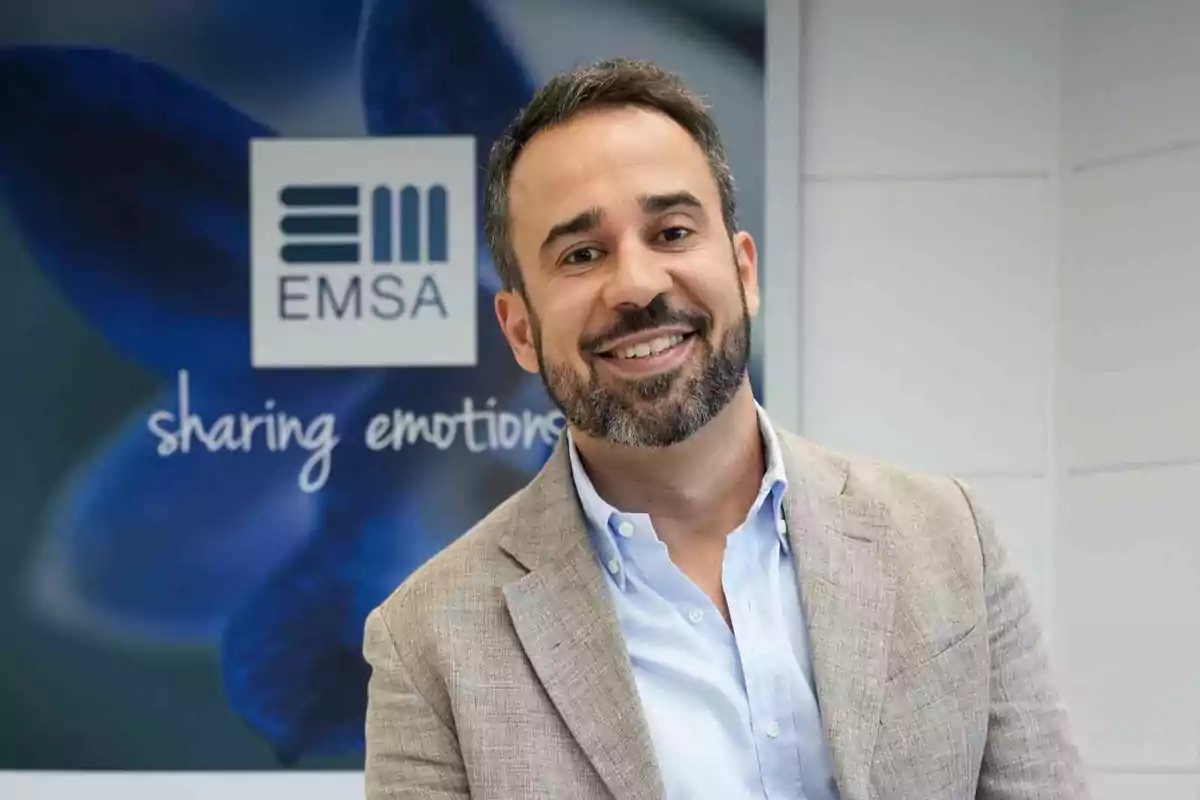 Hombre sonriente con barba y saco claro posando frente a un cartel de EMSA con el texto sharing emotions