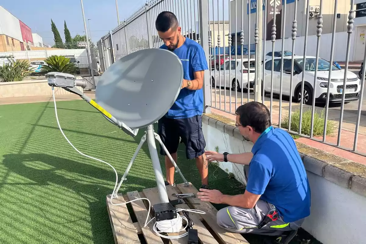 Dos personas instalan una antena parabólica sobre un palé en un área exterior junto a una reja y varios autos estacionados al fondo