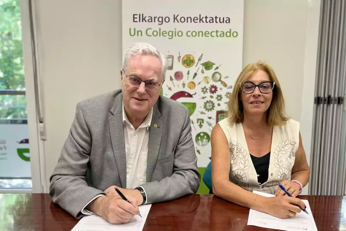 Dos personas sentadas en una mesa firmando documentos con un cartel de fondo que dice Elkargo Konektatua Un Colegio conectado