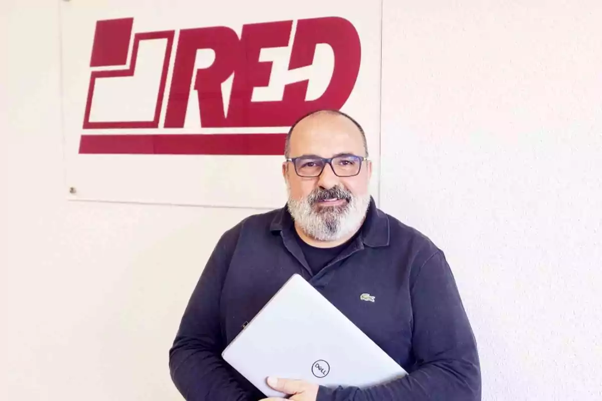 Hombre con barba y gafas sosteniendo una laptop frente a un letrero de la empresa RED