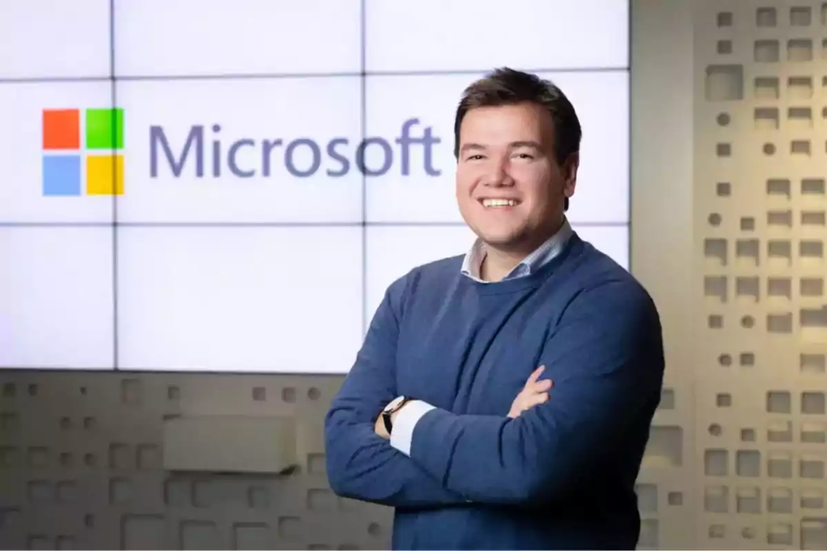 Hombre sonriente con suéter azul de pie frente a un logotipo de Microsoft en una oficina moderna