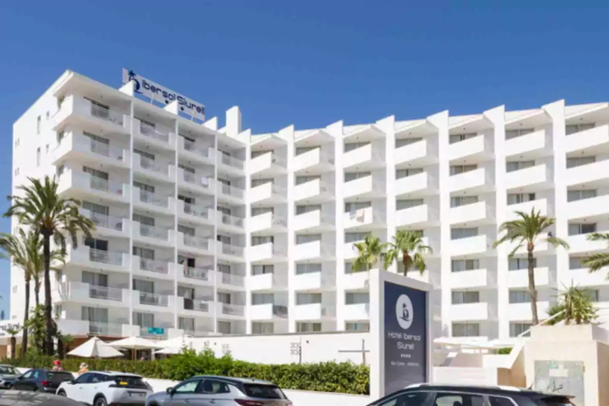 Fachada de un hotel blanco de varios pisos con balcones, palmeras y autos estacionados al frente en un día soleado
