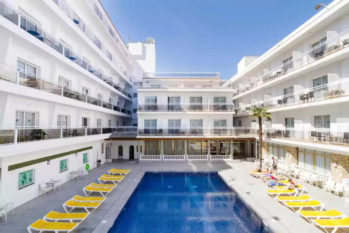 Hotel moderno de varios pisos con balcones y una piscina central rodeada de tumbonas amarillas bajo un cielo despejado