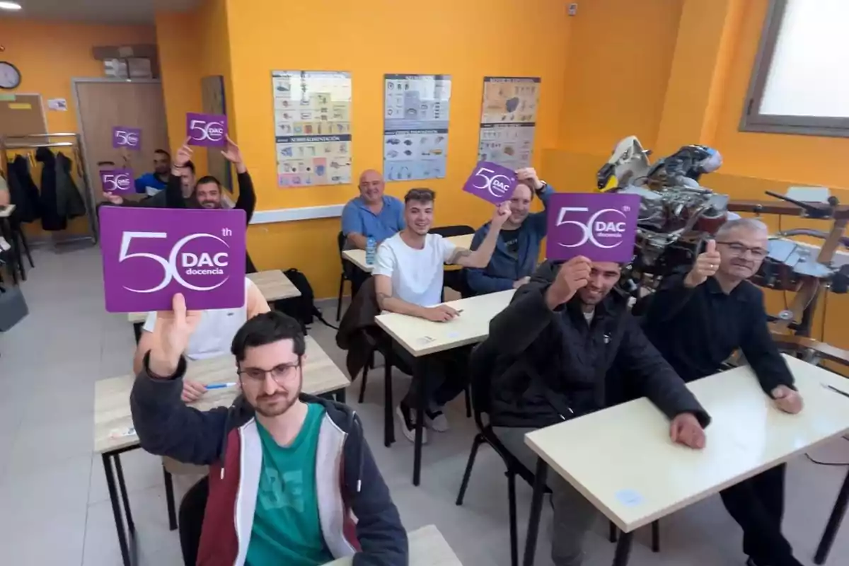 Un grupo de personas en un aula sonríen y levantan carteles morados que celebran el 50 aniversario de DAC docencia