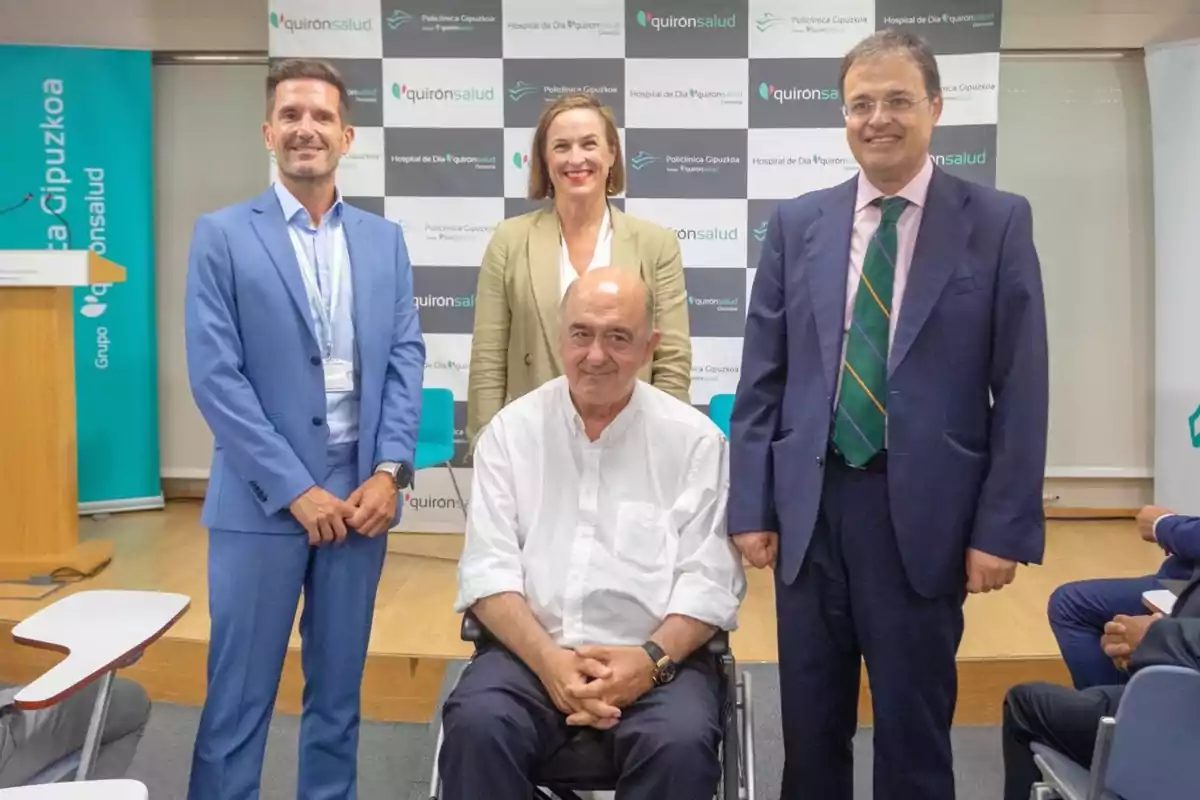 Cuatro personas posan para una foto en un evento corporativo de Quirónsalud, una de ellas está sentada en una silla de ruedas y las otras tres de pie detrás de él, todos sonríen y visten ropa formal.
