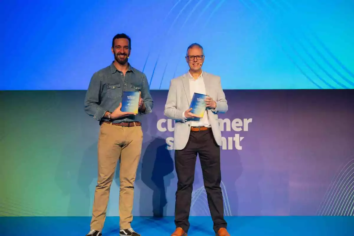 Dos hombres sonrientes posan en un escenario con fondo azul y púrpura mientras sostienen un reconocimiento en las manos
