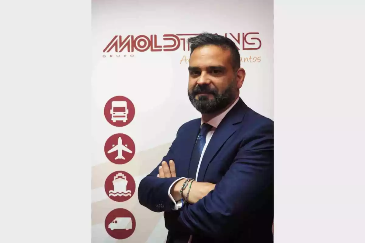 Hombre de barba y traje azul posando con los brazos cruzados frente a un cartel de empresa de transporte con iconos de camión, avión, barco y furgoneta