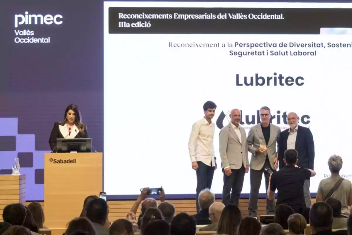 Un grupo de personas en un escenario recibe un reconocimiento empresarial mientras una mujer presenta el evento desde un atril con el logo de Sabadell y pimec, frente a una audiencia que toma fotos