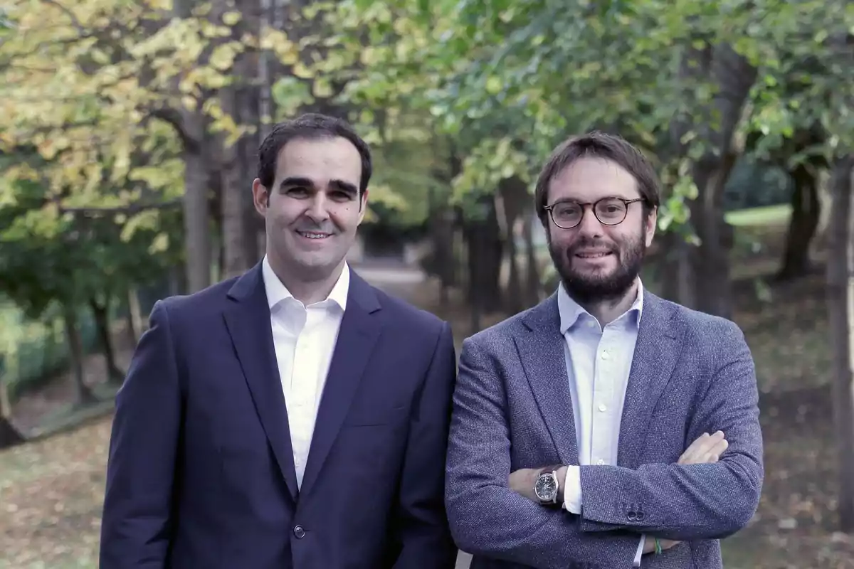 Dos hombres con ropa formal posan sonrientes en un parque con árboles al fondo