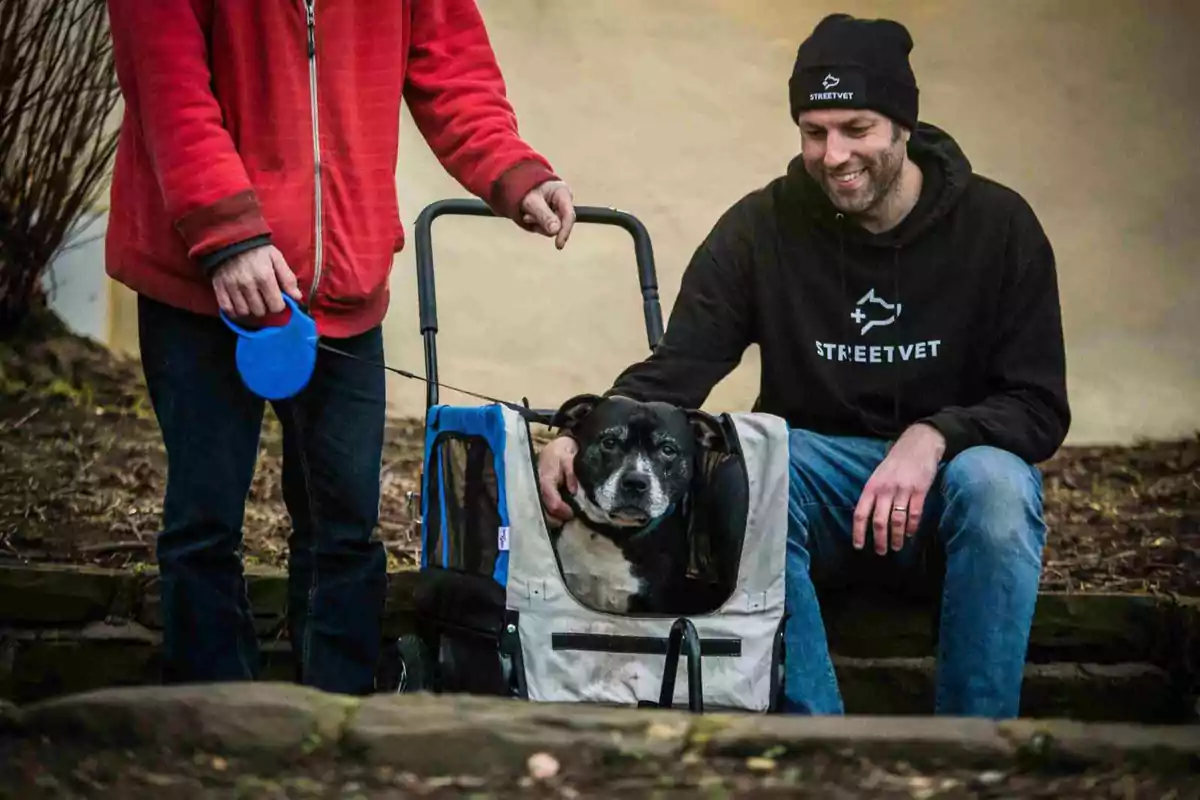 Un perro negro y blanco está sentado en un carrito mientras un hombre con sudadera y gorro negro lo acaricia y otra persona de pie sostiene la correa azul del perro.