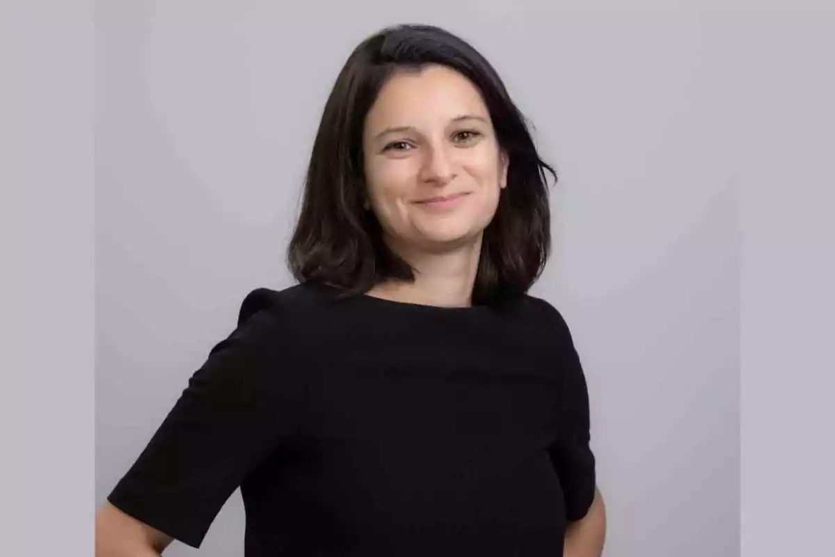 Mujer de cabello oscuro y liso con una ligera sonrisa, vestida con blusa negra y fondo gris claro