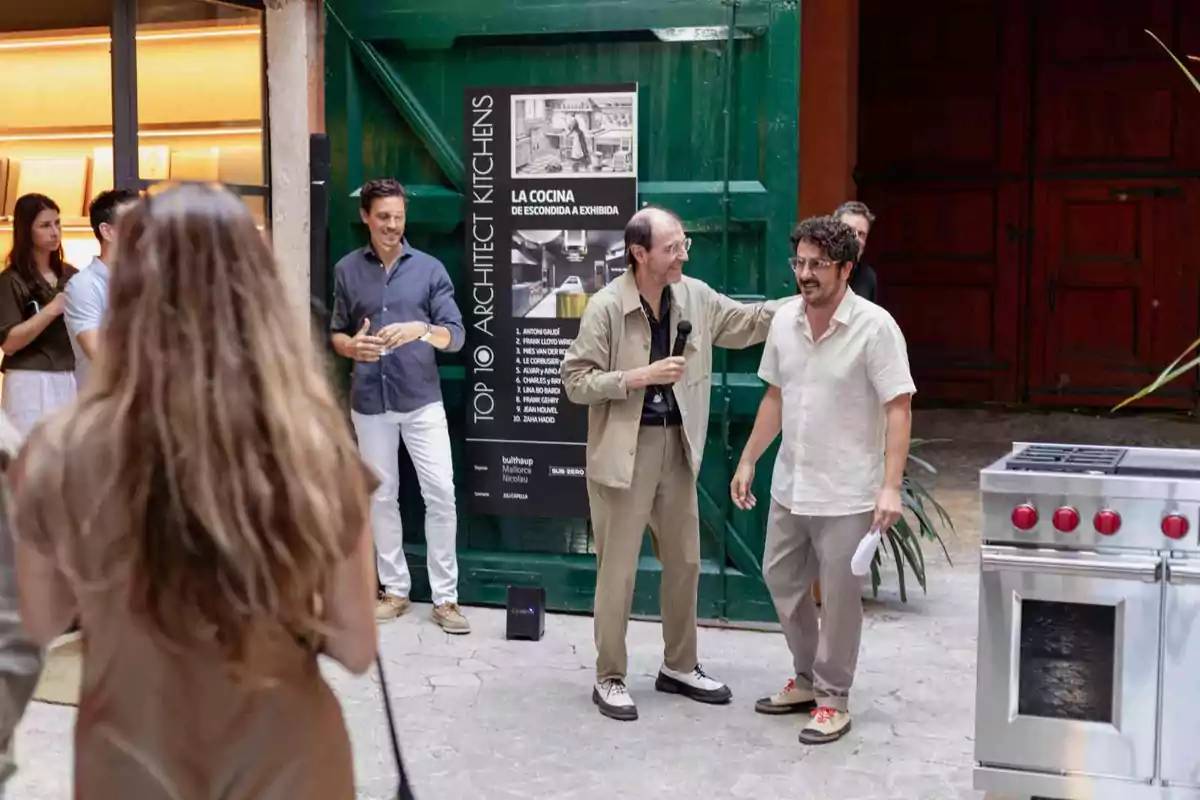 Un grupo de personas participa en un evento al aire libre frente a un cartel informativo y una estufa plateada, mientras dos hombres en el centro interactúan con un micrófono y el público observa.