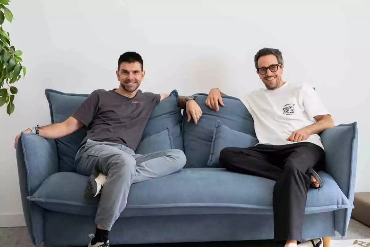 Dos hombres sonrientes sentados en un sofá azul en una habitación luminosa