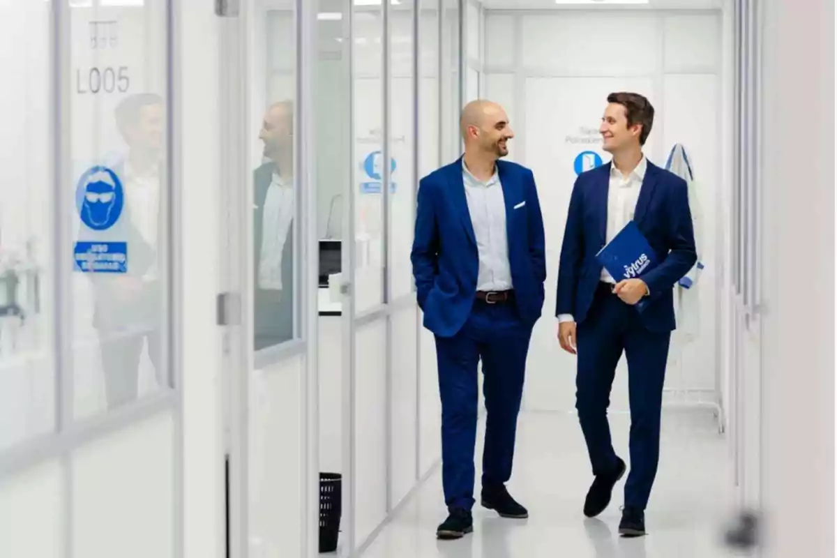 Dos hombres con traje azul caminan y conversan sonrientes por un pasillo de oficinas modernas y luminosas