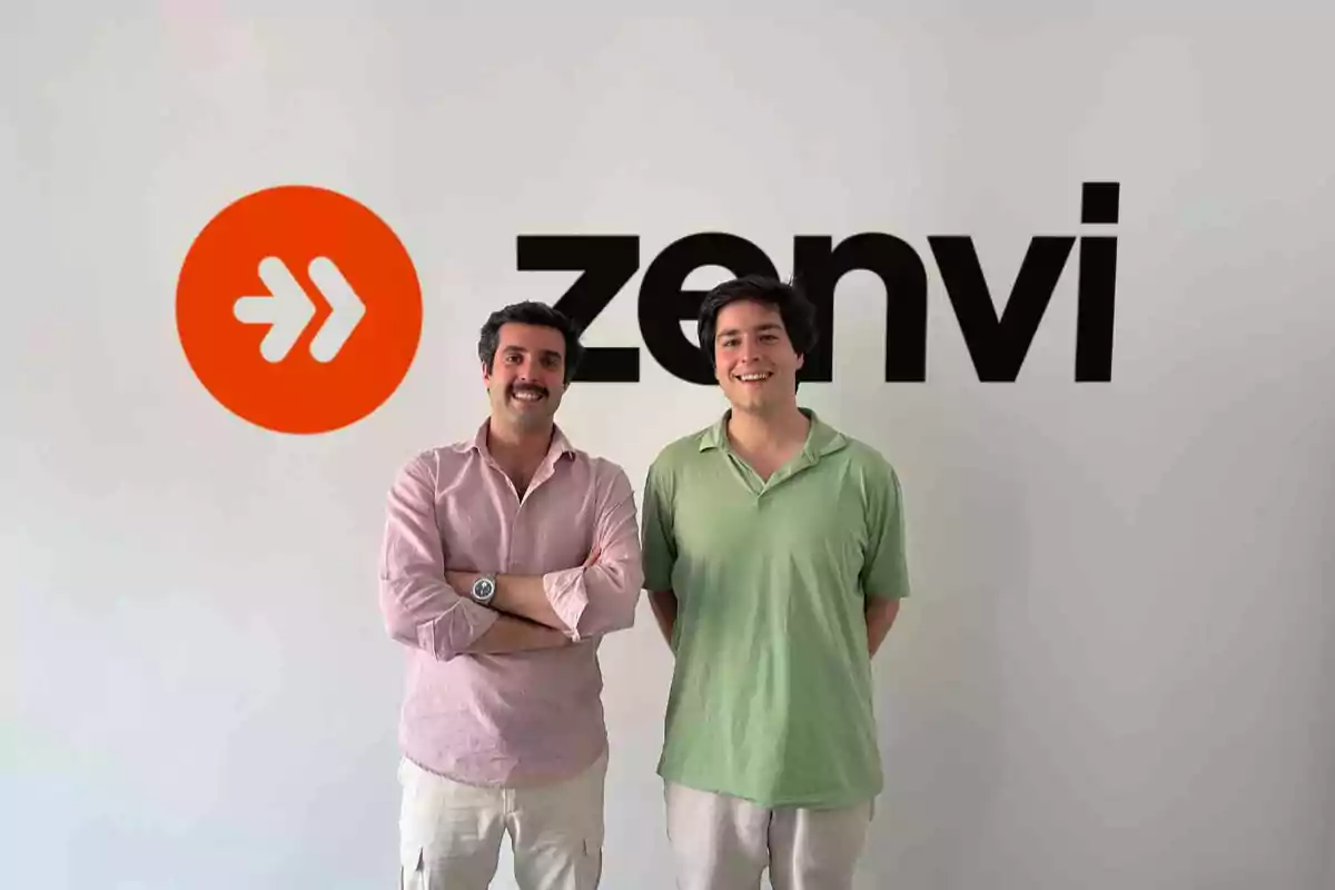 Dos personas posan sonrientes frente a una pared blanca con el logo y el nombre de la empresa Zenvi
