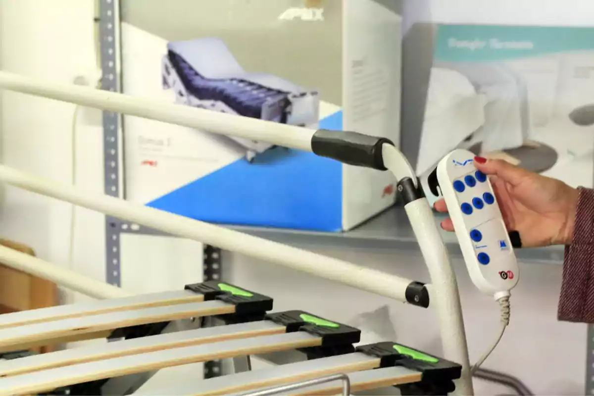 Persona sosteniendo un control remoto de una cama hospitalaria eléctrica junto a una estructura metálica y cajas en el fondo