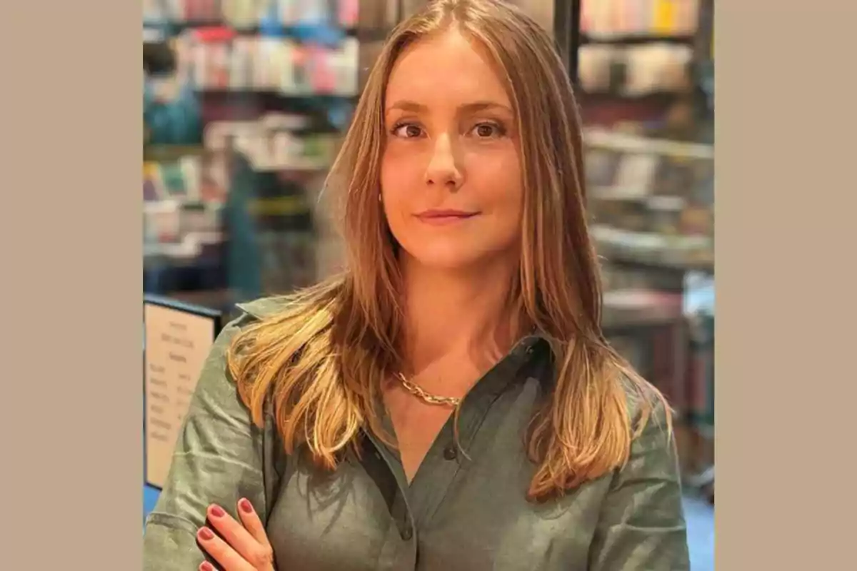 Mujer joven de cabello rubio y lacio con camisa verde posando con los brazos cruzados en un entorno de librería desenfocada
