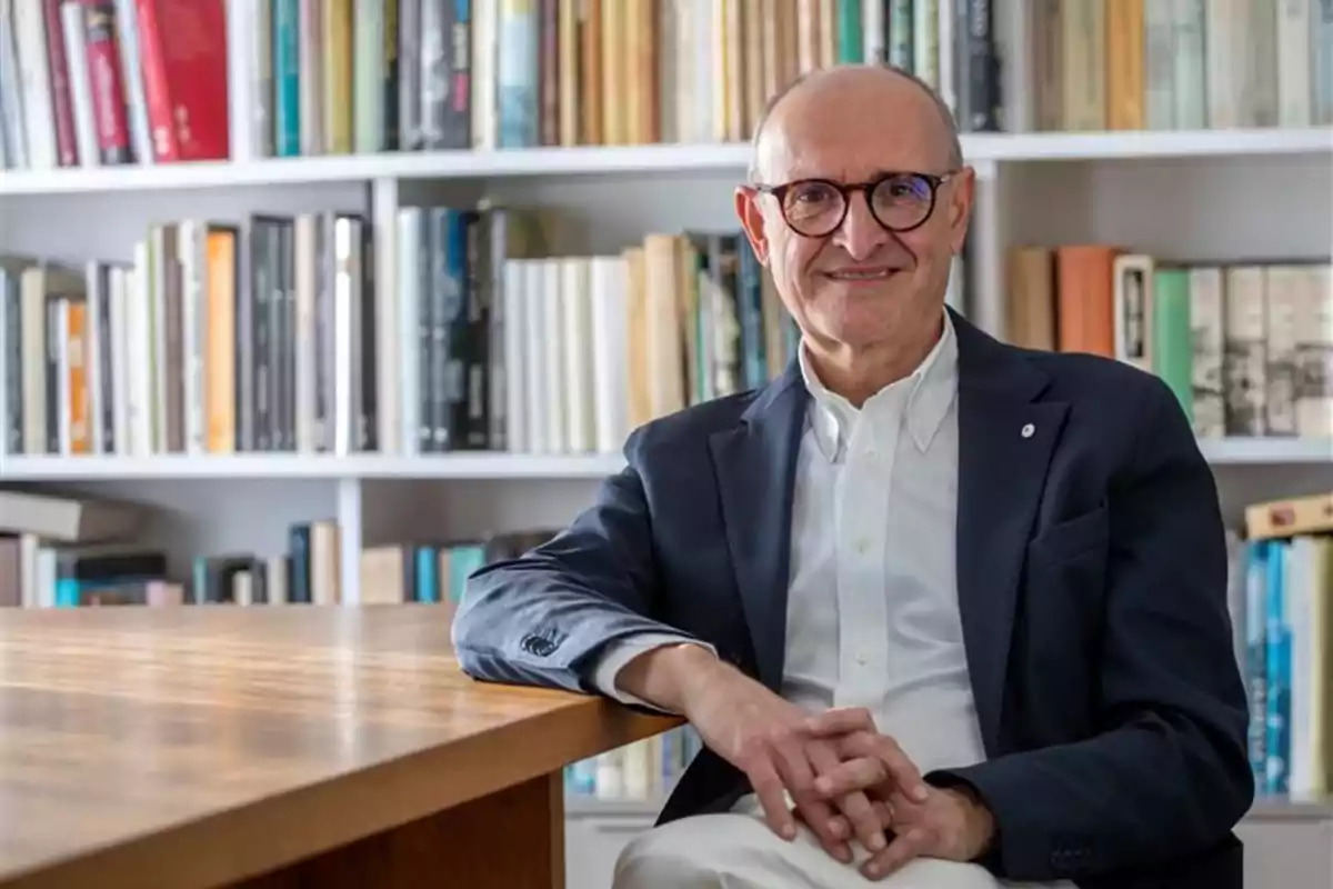 Hombre mayor con gafas y saco oscuro sentado frente a una mesa de madera con estanterías llenas de libros al fondo
