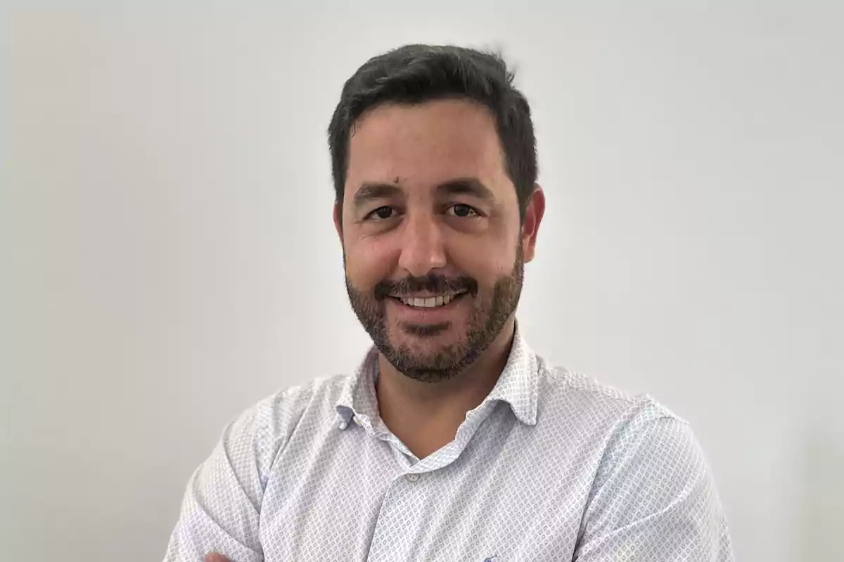 Hombre de cabello corto y barba con camisa clara sonriendo frente a un fondo blanco