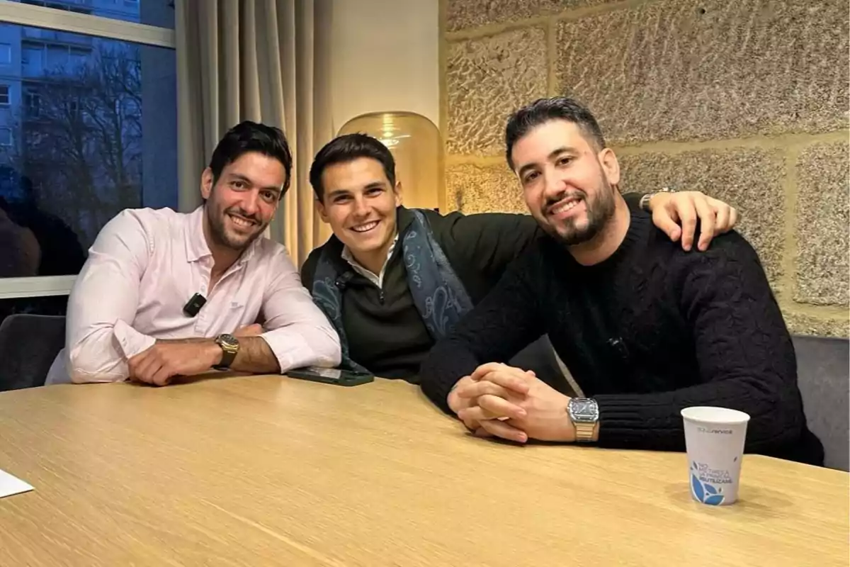 Tres hombres sonríen sentados alrededor de una mesa de madera en una sala iluminada, con una ventana y una pared de piedra al fondo.