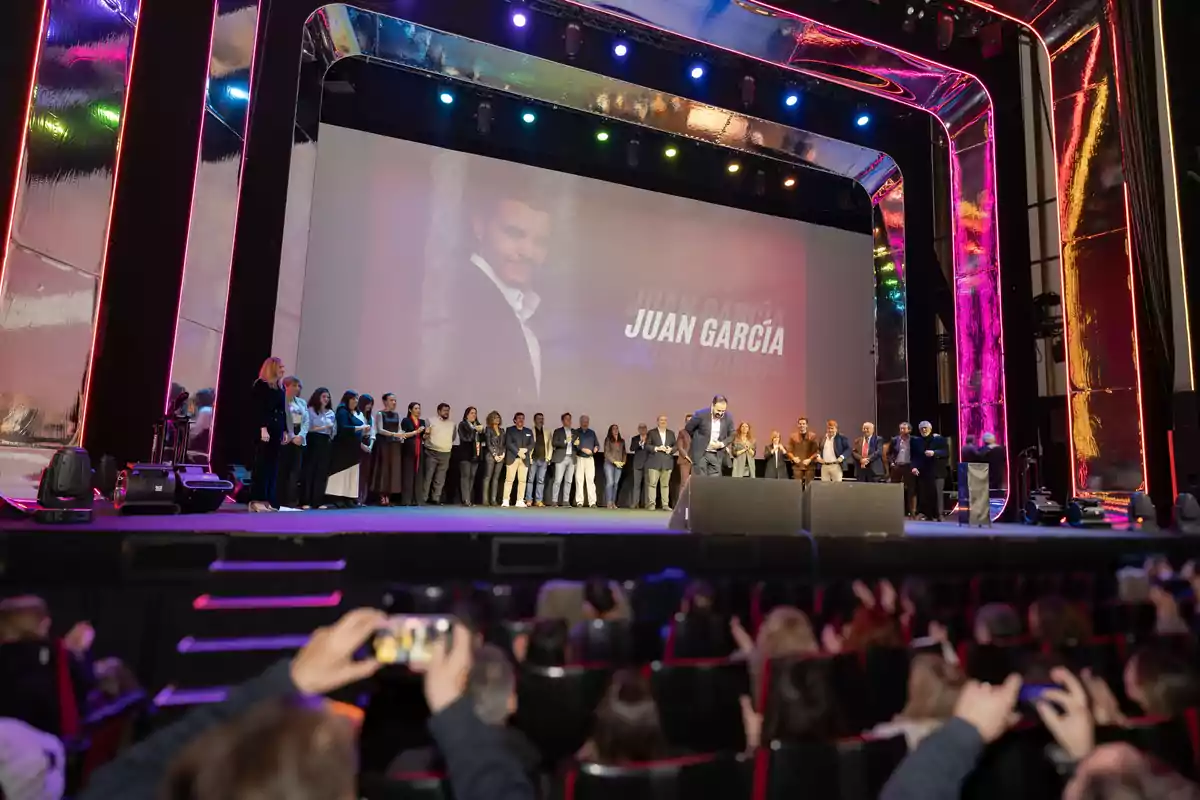 Un grupo de personas está reunido en un escenario iluminado con luces de colores mientras una audiencia observa y toma fotos, en la pantalla se proyecta el nombre Juan García junto a una imagen de un hombre.