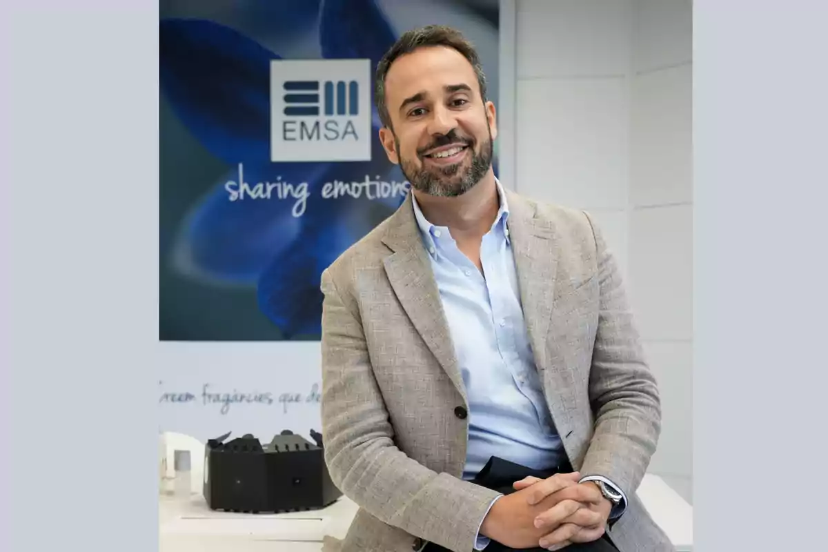 Hombre sonriente con barba y saco claro sentado en una oficina moderna con un cartel de fondo que dice EMSA sharing emotions