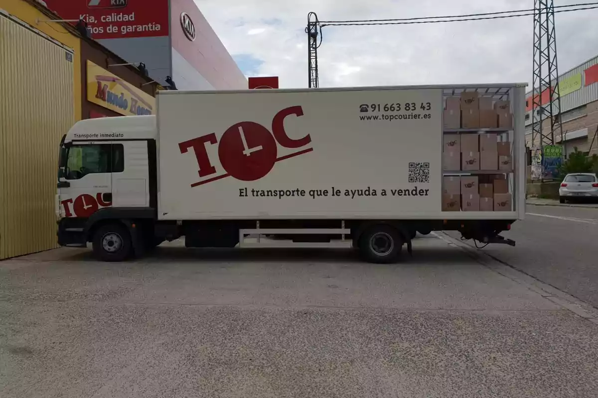 Camión de reparto de la empresa TOC estacionado junto a una nave industrial con cajas apiladas impresas en el lateral del vehículo y el lema El transporte que le ayuda a vender