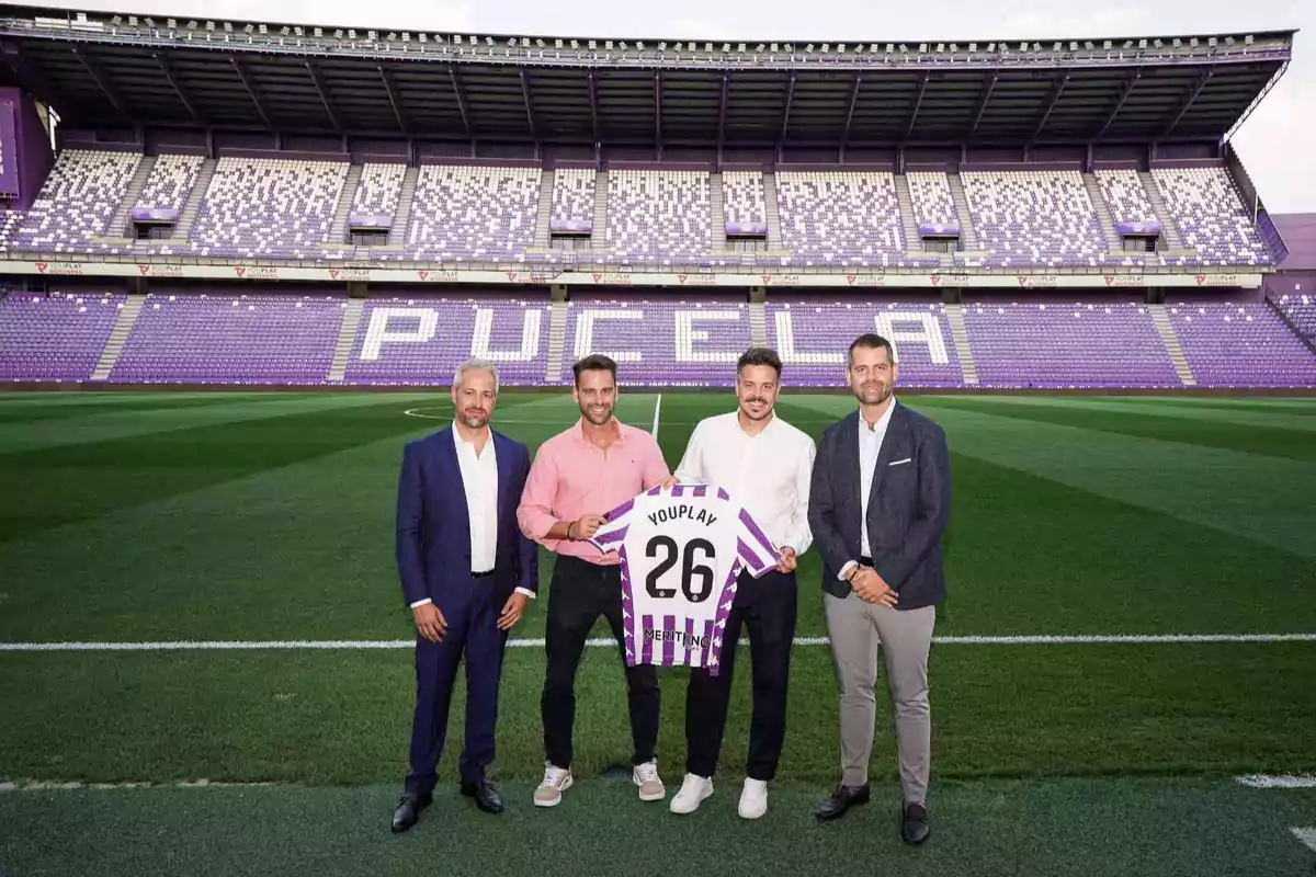 Cuatro hombres posan en el campo de un estadio de fútbol con gradas moradas y la palabra Pucela al fondo mientras dos de ellos sostienen una camiseta blanca y morada con el número 26 y el nombre Youplay