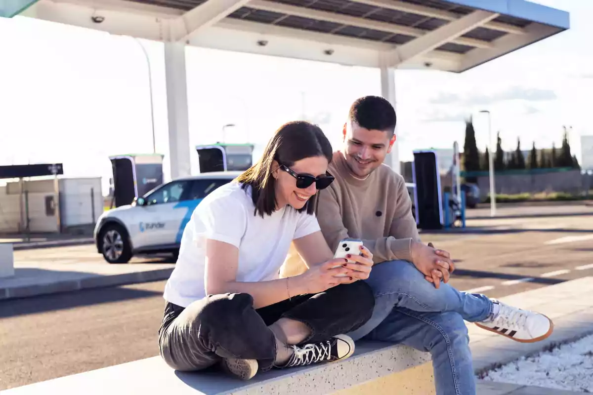 Dos personas jóvenes sonríen mientras miran un teléfono móvil sentados en una estación de carga para autos eléctricos