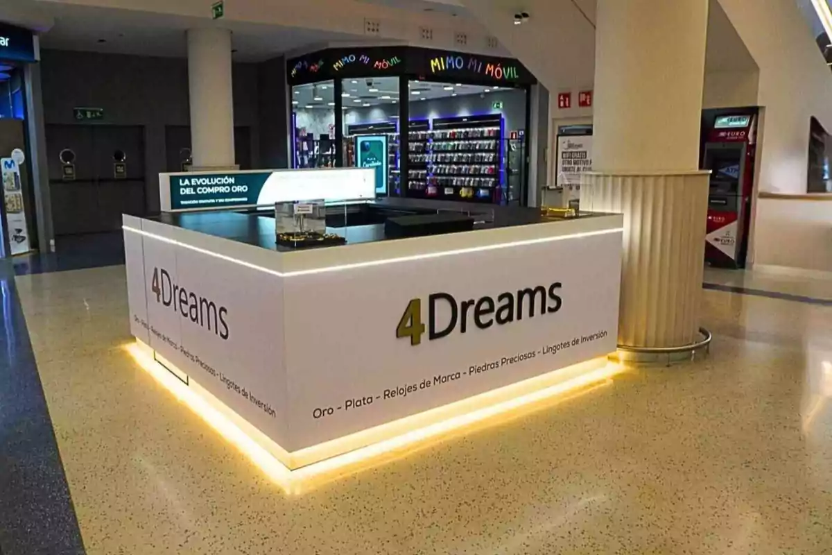 Puesto de compra y venta de oro y relojes de marca llamado 4Dreams en el interior de un centro comercial con iluminación en la base y una tienda de telefonía al fondo