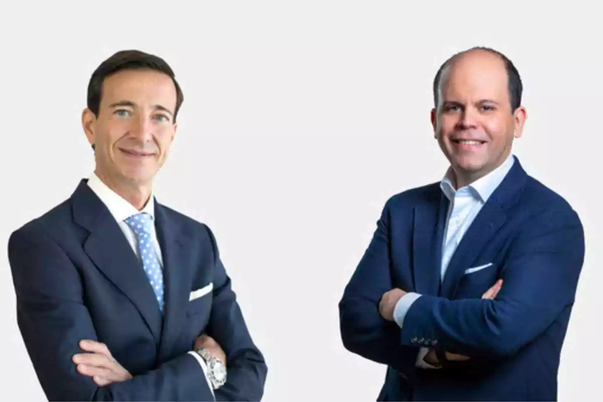 Dos hombres de negocios con traje posan con los brazos cruzados y sonríen frente a un fondo claro